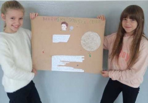 Dwie uczennice klasy 3b trzymają wykonany przez siebie plakat przedstawiający postać Marii Skłodowskiej-Curie.