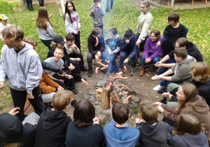 Uczniowie siedzą w kręgu trzymając kiełbaski nad ogniskiem.