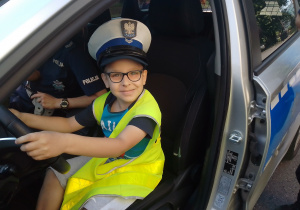 Uczeń w czapce i kamizelce policyjnej siedzi za kierownicą radiowozu.