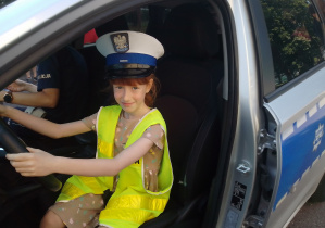 Uczennica w czapce i kamizelce policyjnej siedzi za kierownicą radiowozu.