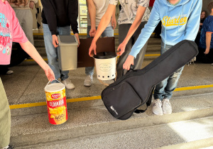Uczniowie, bez twarzy, pokazują pokrowiec do gitary, pudełko po chipsach, starą walizkę i kosz na śmieci.