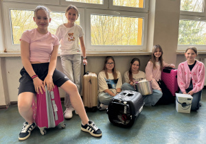Sześć uczennic pokazuje swoje pomysły na plecak szkolny: walizki podróżne, transporter dla kota, mini-pralkę i garnek.