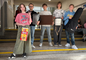 Pięcioro uczniów stoi na schodach z koszem na śmieci, pokrowcem na gitarę, walizką oraz dużym opakowaniem po chipsach zamiast plecaka.