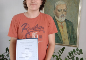 Laureat Wojewódzkiego Konkursu Przedmiotowego z Języka Angielskiego z dyplomem, na tle reprodukcji przedstawiającej patrona szkoły