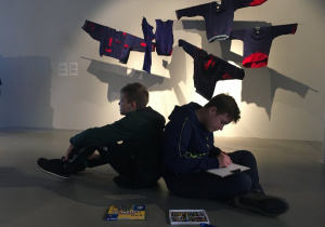 Chłopcy siedzą tyłem do siebie na tle dekoracji jaką stanowią zawieszone pod sufitem kolorowe bluzy.